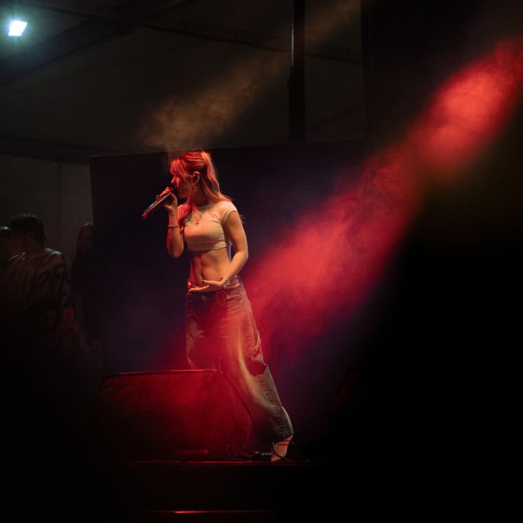 Una artista está de pie en el escenario con un micrófono en la mano, rodeada de luces rojas y moradas y efectos de humo. Lleva un top blanco ajustado y unos vaqueros holgados, concentrada en su actuación mientras se ve al público al fondo.